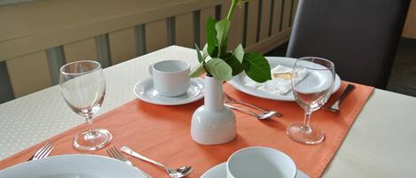 Una mesa de comedor con platos blancos, tazas y cubiertos, un florero con una sola rosa naranja y una ventana con vista a un edificio.