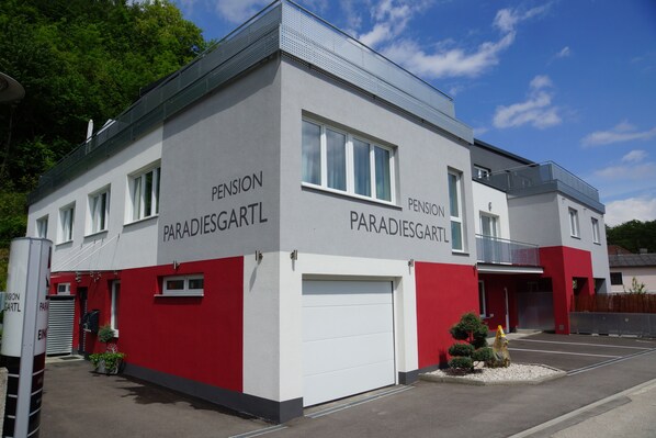 A building with the name 'Pension Paradiesgartl' on its facade, featuring a white and red color scheme, a garage door, and a small garden area with a fire hydrant. - Pension Paradiesgartl (Amstetten)