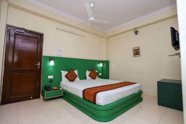 Une chambre d’hôtel avec une tête de lit verte, un lit avec une literie blanche et des oreillers marron, une table de chevet avec un téléphone et un chargeur, un ventilateur de plafond et une armoire verte.