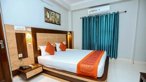 Una habitación de hotel con cabecera de madera, cama con ropa de cama blanca y detalles naranja, mesita de noche con teléfono y lámpara, y cortinas color turquesa.
