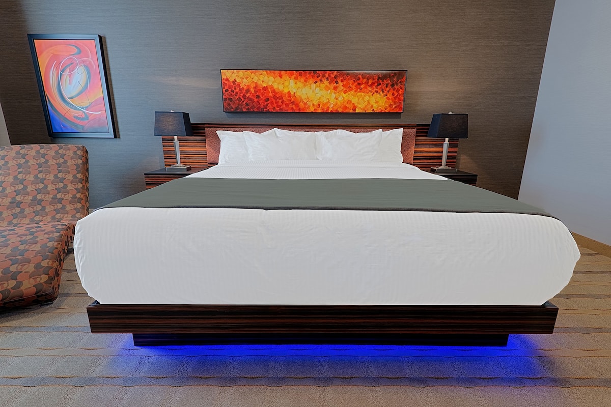 Una habitación de hotel moderna con una cama grande, dos mesitas de noche, un televisor montado en la pared y un sillón estampado.