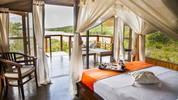 A bedroom with a canopy bed, a wooden chair, a table with a tray, and a view of greenery outside. - Naara Ecolodge & Spa (Chidenguele)