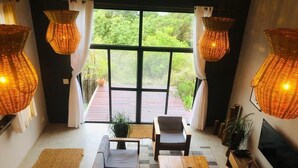 A modern living room with a sofa, coffee table, and TV. There are wicker lamps and a large window with a view of greenery. - Naara Ecolodge & Spa (Chidenguele)