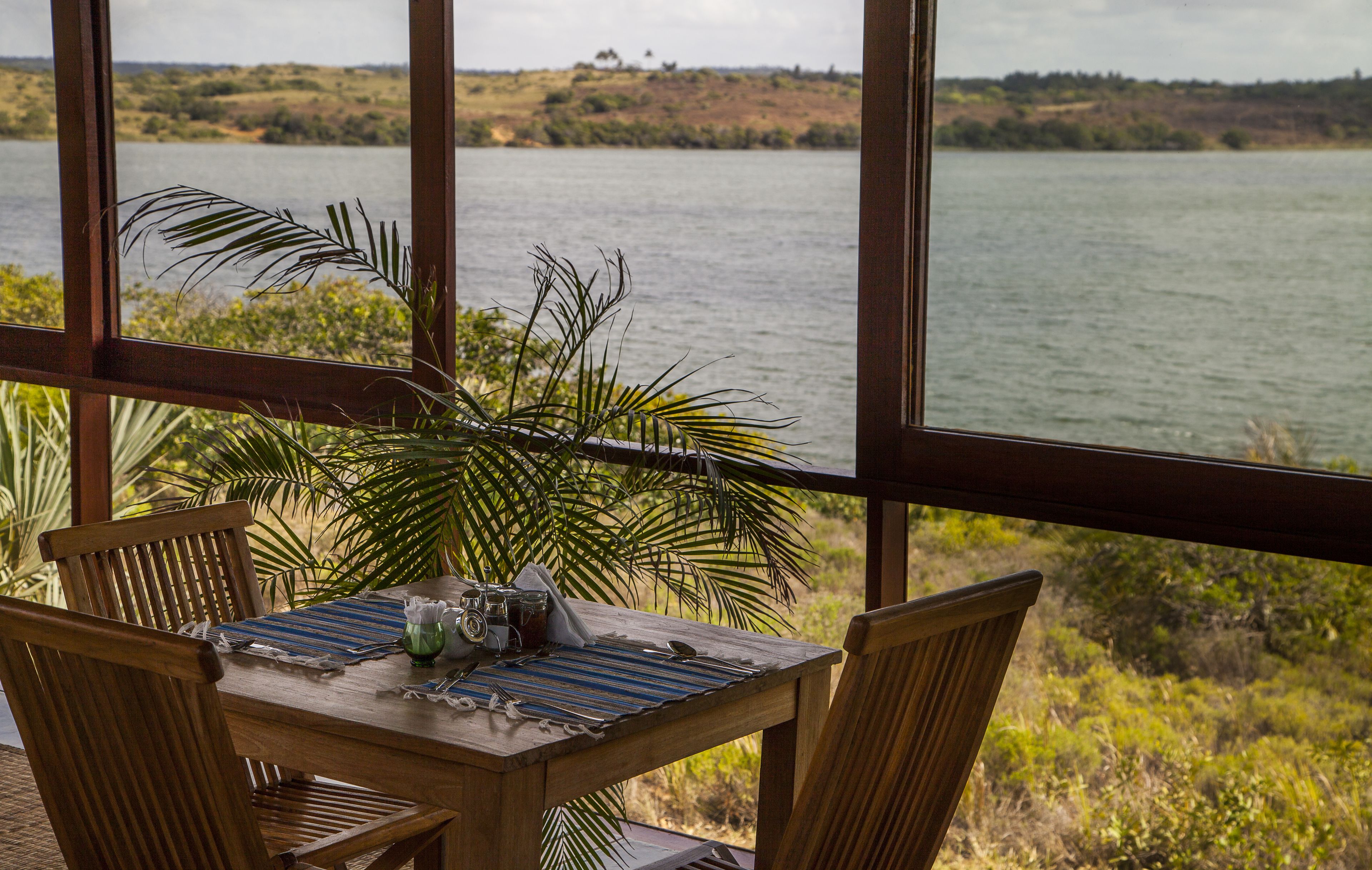 A wooden table with chairs and a camera, overlooking a lake with vegetation and hills in the background.