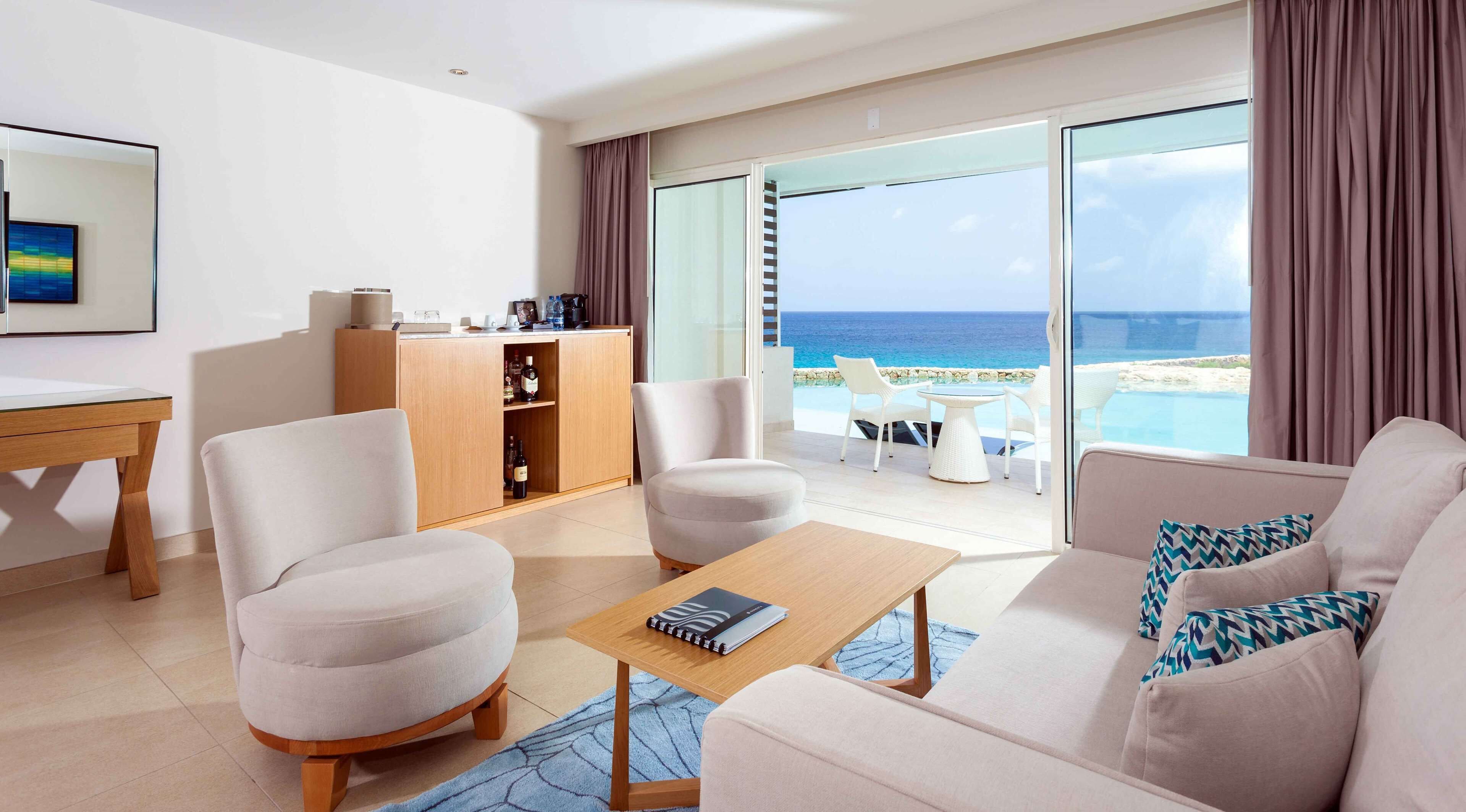 Una habitación de hotel moderna con sofá, sillones, una mesita de madera, minibar y un balcón con vista al océano.