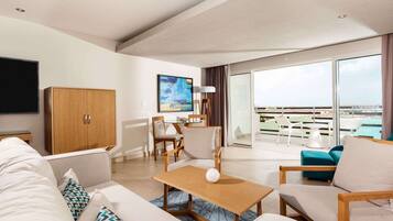A modern hotel room with a large flat-screen TV, a wooden cabinet, a dining area with chairs, a sofa, and a view of the city through large windows.