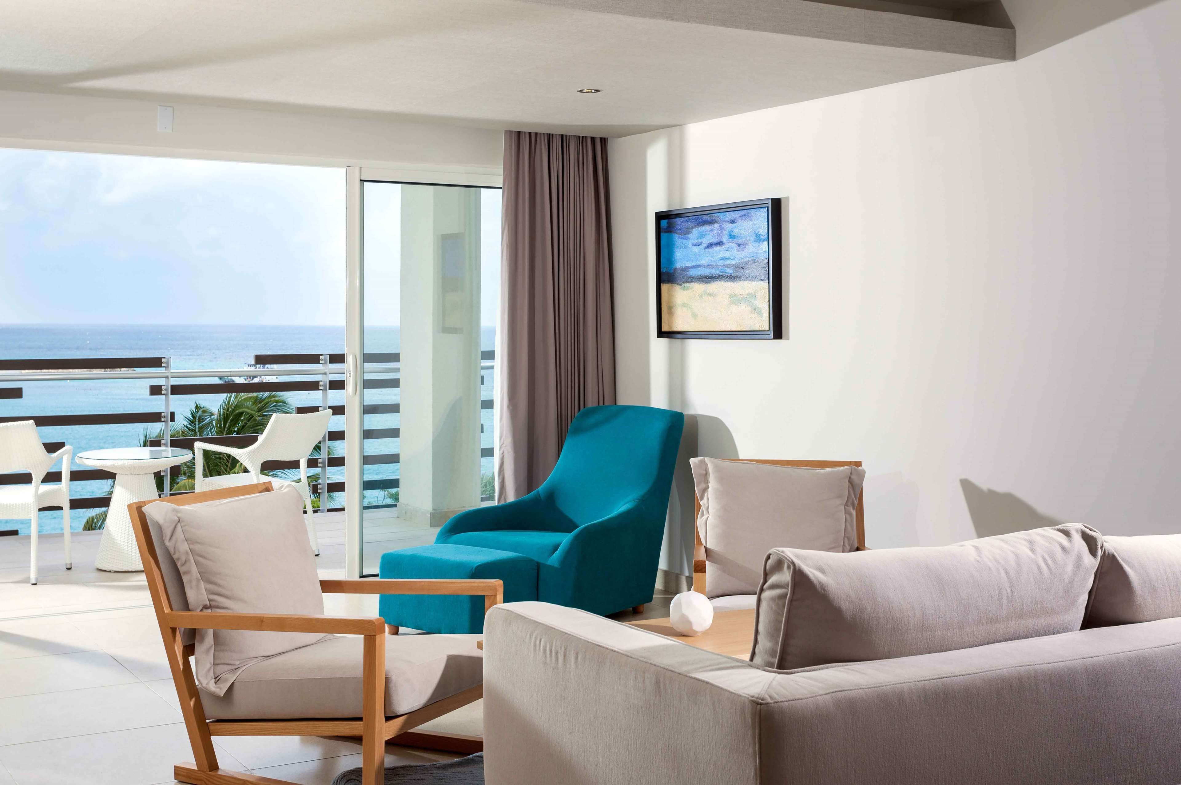 Habitación de hotel moderna con balcón y vistas al mar, que incluye un sofá, sillones y una mesita.