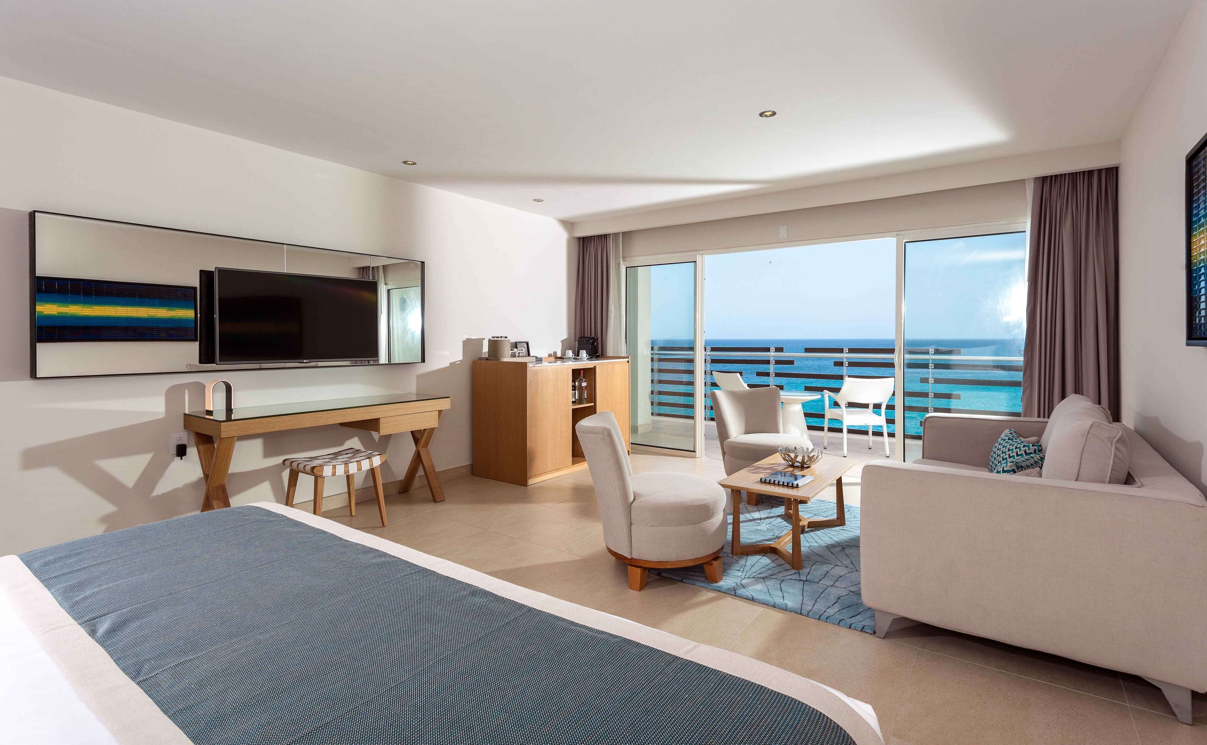 Habitación de hotel moderna con una cama grande, un escritorio con computadora, un área de descanso con vista al océano y un balcón con puerta de vidrio.