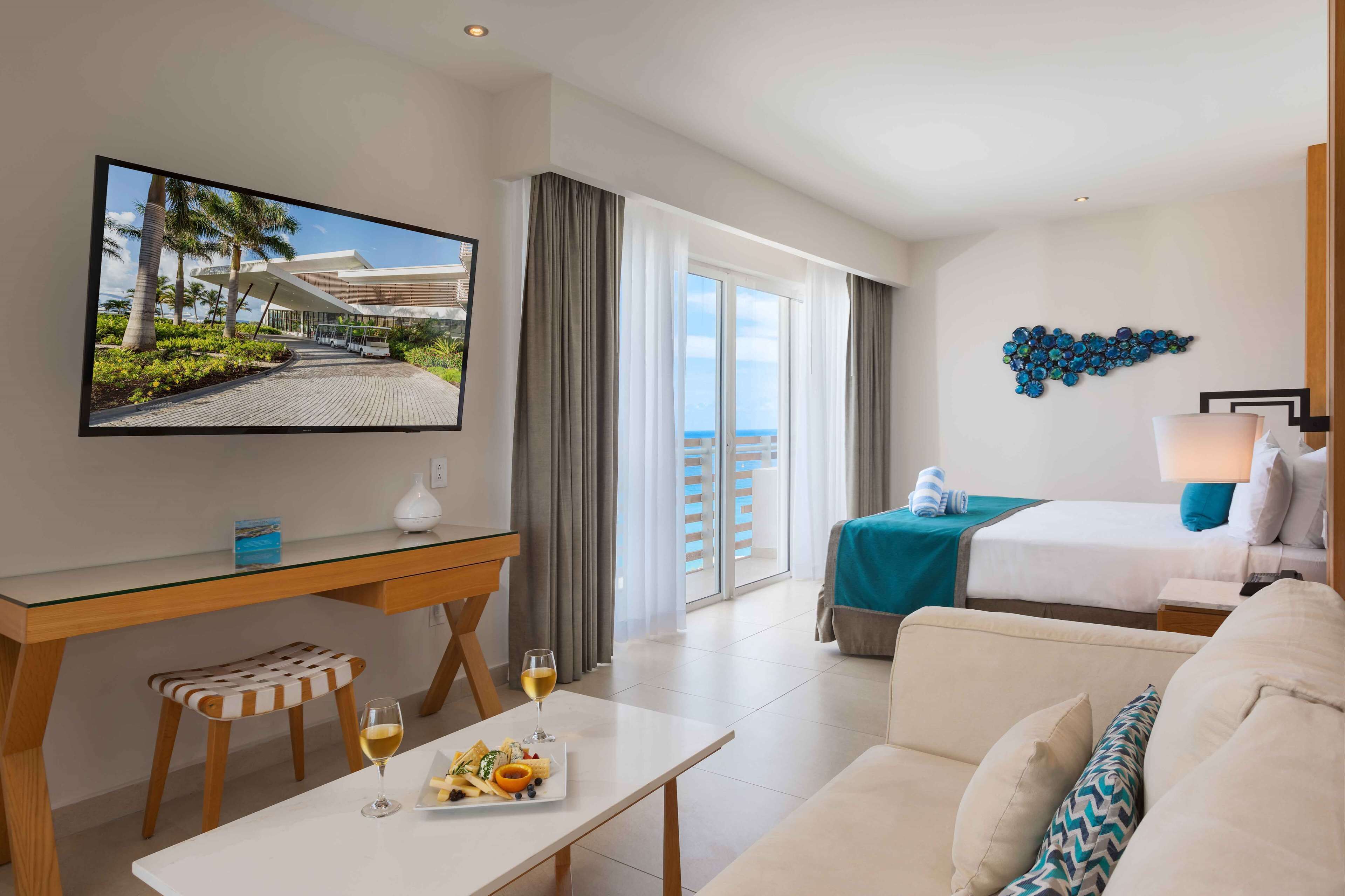 A hotel room with a bed, a TV, a dining table with a plate of food and a glass of wine, a sofa, and a balcony with a view of a building and palm trees.