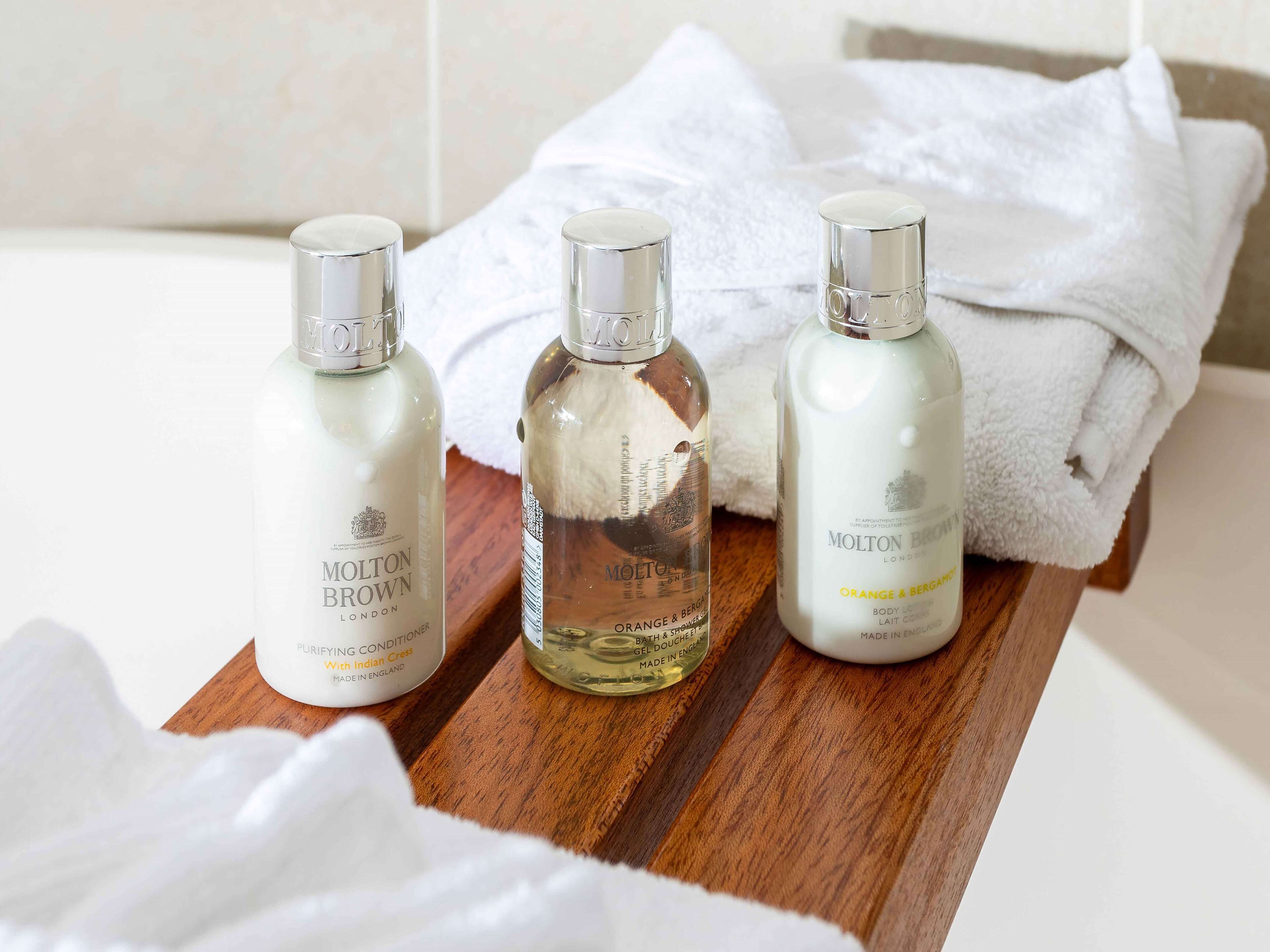 Trois flacons de lotion pour le corps Molton Brown London, présentés sur un plateau en bois, avec des serviettes blanches en arrière-plan.