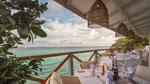 A dining table set with fine dining utensils and a candle, overlooking a turquoise sea with a sailboat in the distance.