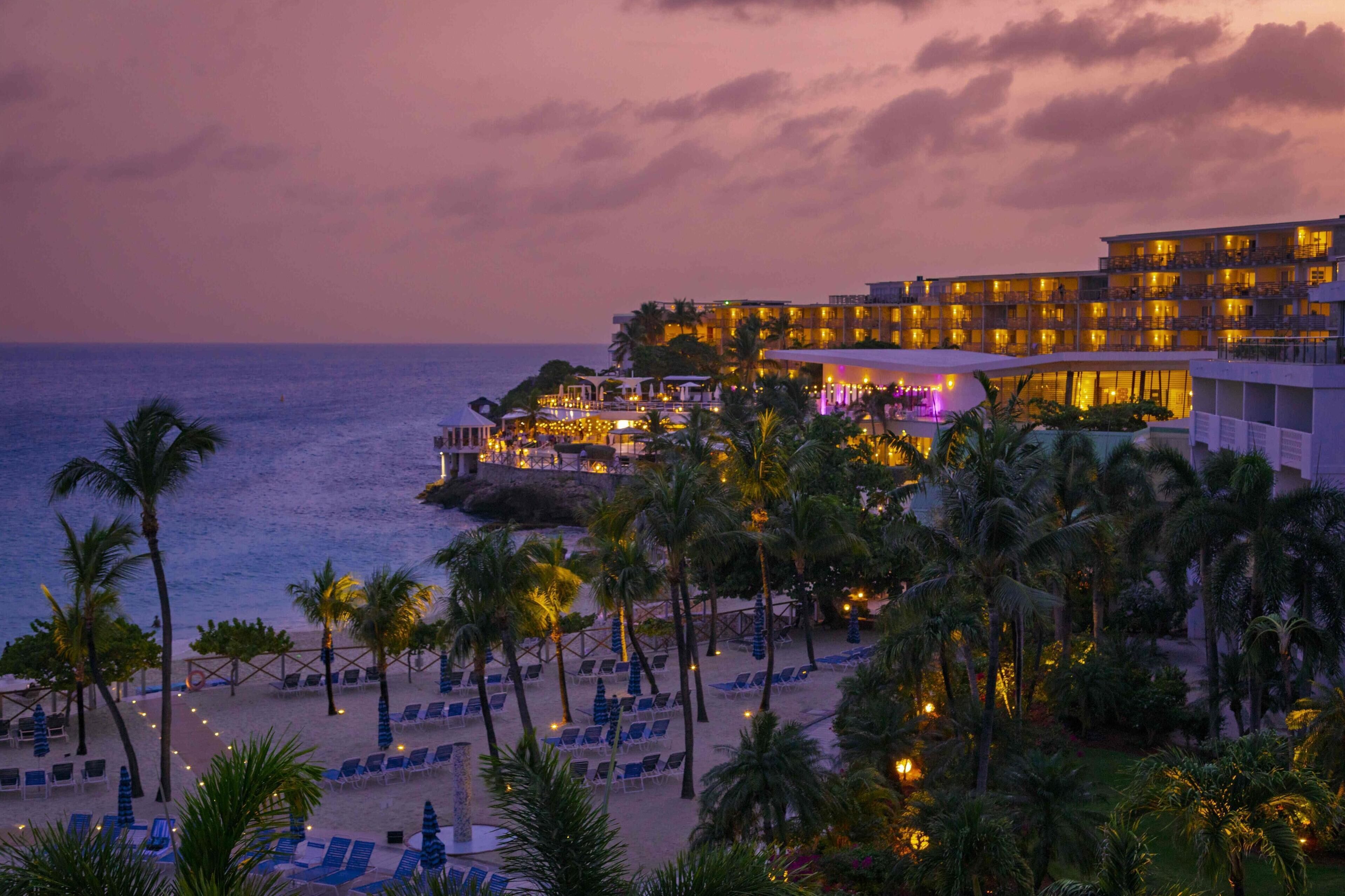 Un hotel frente a la playa con varios balcones, palmeras y sillas de playa.
