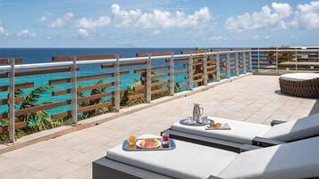 A rooftop terrace with a view of the ocean, outdoor seating with cushions, and a breakfast setup.
