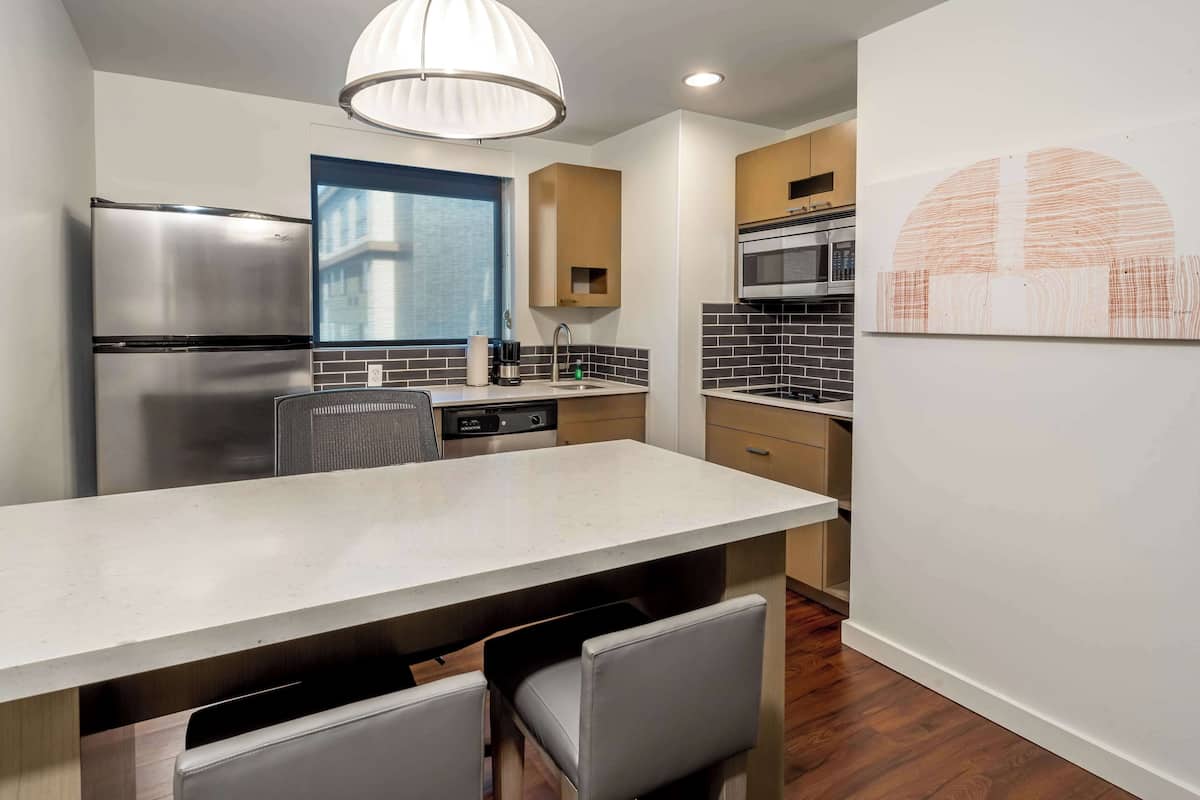 A modern kitchen with a stainless steel refrigerator, a built-in dishwasher, a large island with seating, and a hanging light fixture.