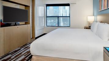 A hotel room with a large bed, a flat-screen TV, a window with a city view, and a bedside table with a lamp.