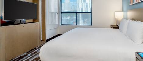 A hotel room with a large bed, a flat-screen TV, a window with a city view, and a bedside table with a lamp.