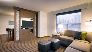 A hotel room with a sectional sofa, ottomans, a flat-screen TV, a desk with a lamp, and a large window with a city view. - Hyatt House Salt Lake City Downtown (Salt Lake City)