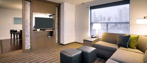 A hotel room with a sectional sofa, ottomans, a flat-screen TV, a desk with a lamp, and a large window with a city view.