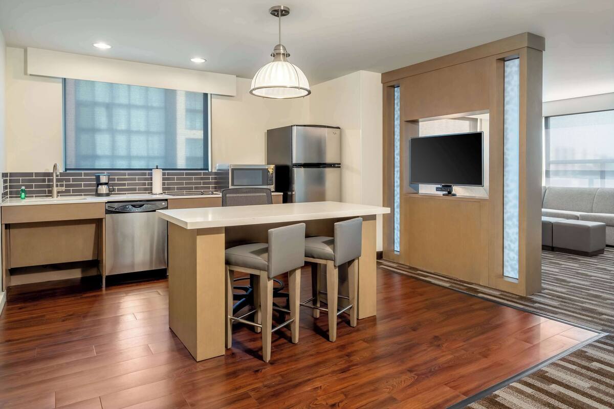 A modern kitchen with a central island, bar stools, a refrigerator, a microwave, and a flat-screen TV.
