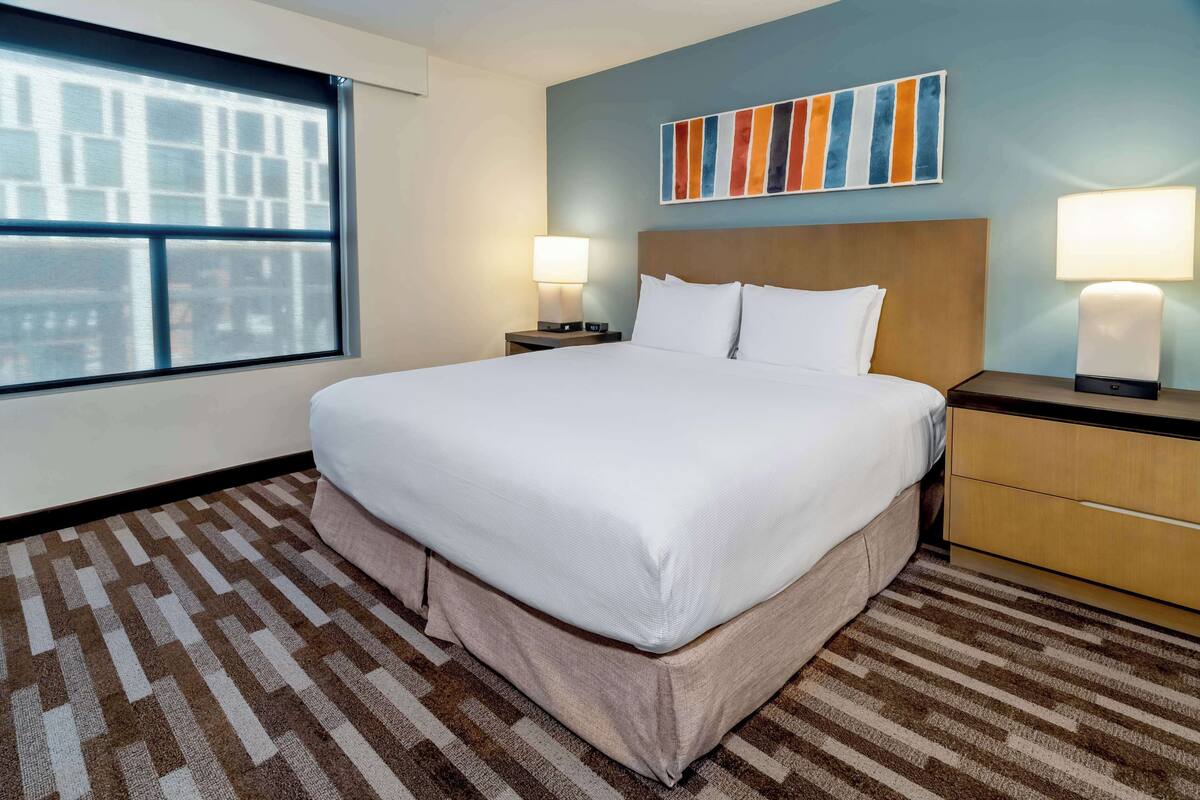 A hotel room with a large bed, two bedside tables with lamps, a window with a view of buildings, and a colorful abstract painting on the wall.