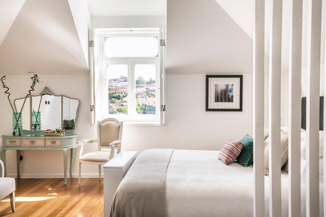 A bedroom with a bed, a desk, a chair, a window with a view, and a framed picture on the wall.