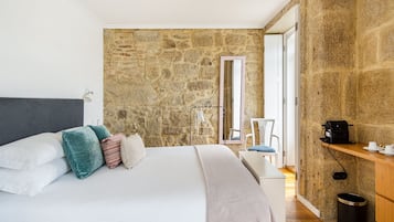 Un dormitorio con pared de piedra, una cama con ropa de cama blanca, una mesita de noche con cafetera y una silla.