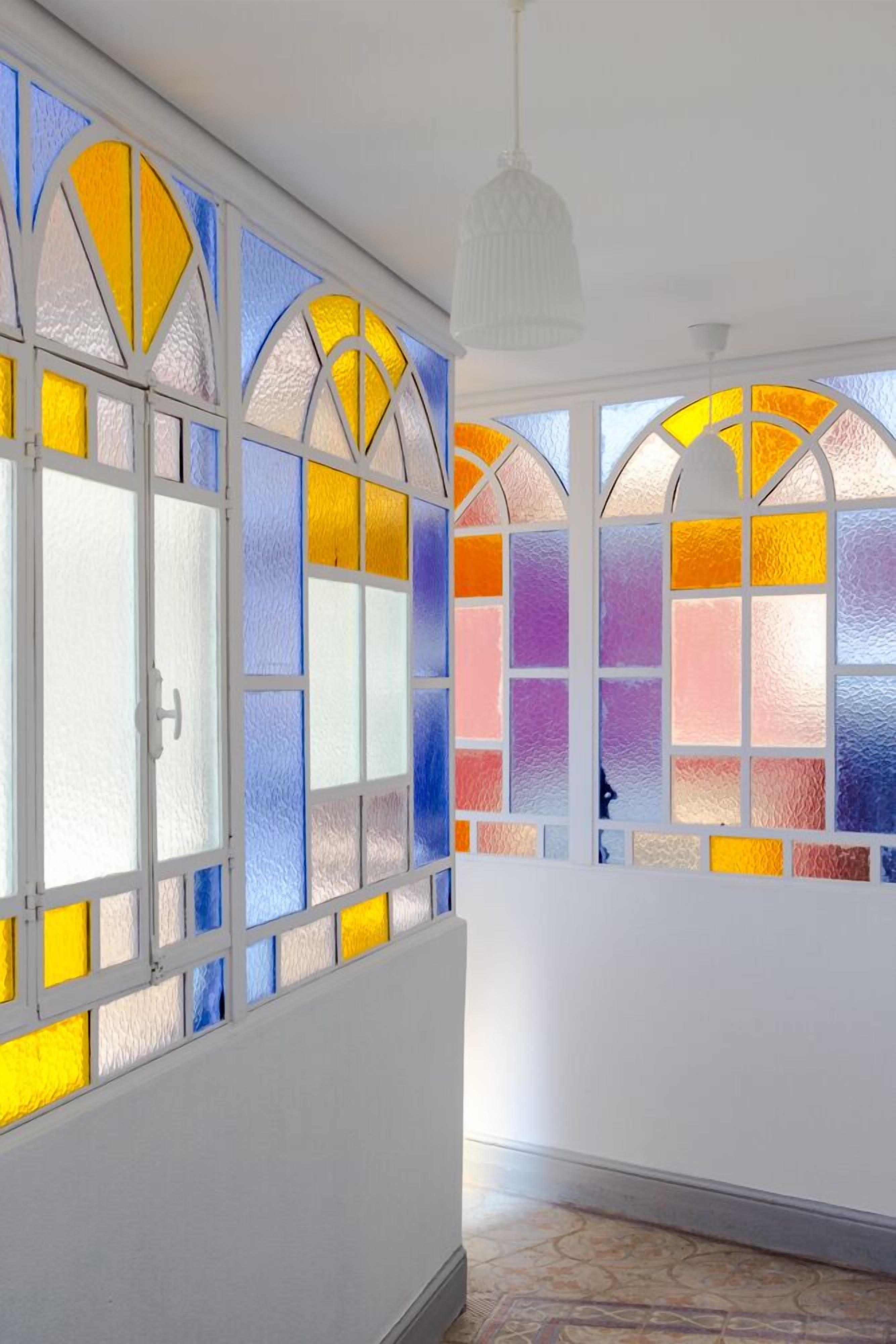 Una habitación con amplios ventanales con vidrieras de colores, paredes blancas y una lámpara colgante.