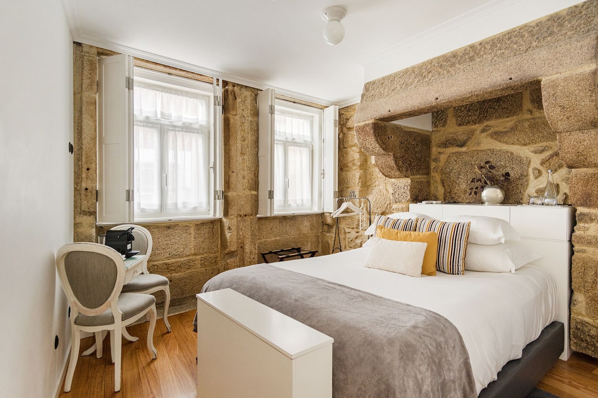 A bedroom with a stone wall, a bed with multiple pillows, a desk with a chair, and a window with curtains.