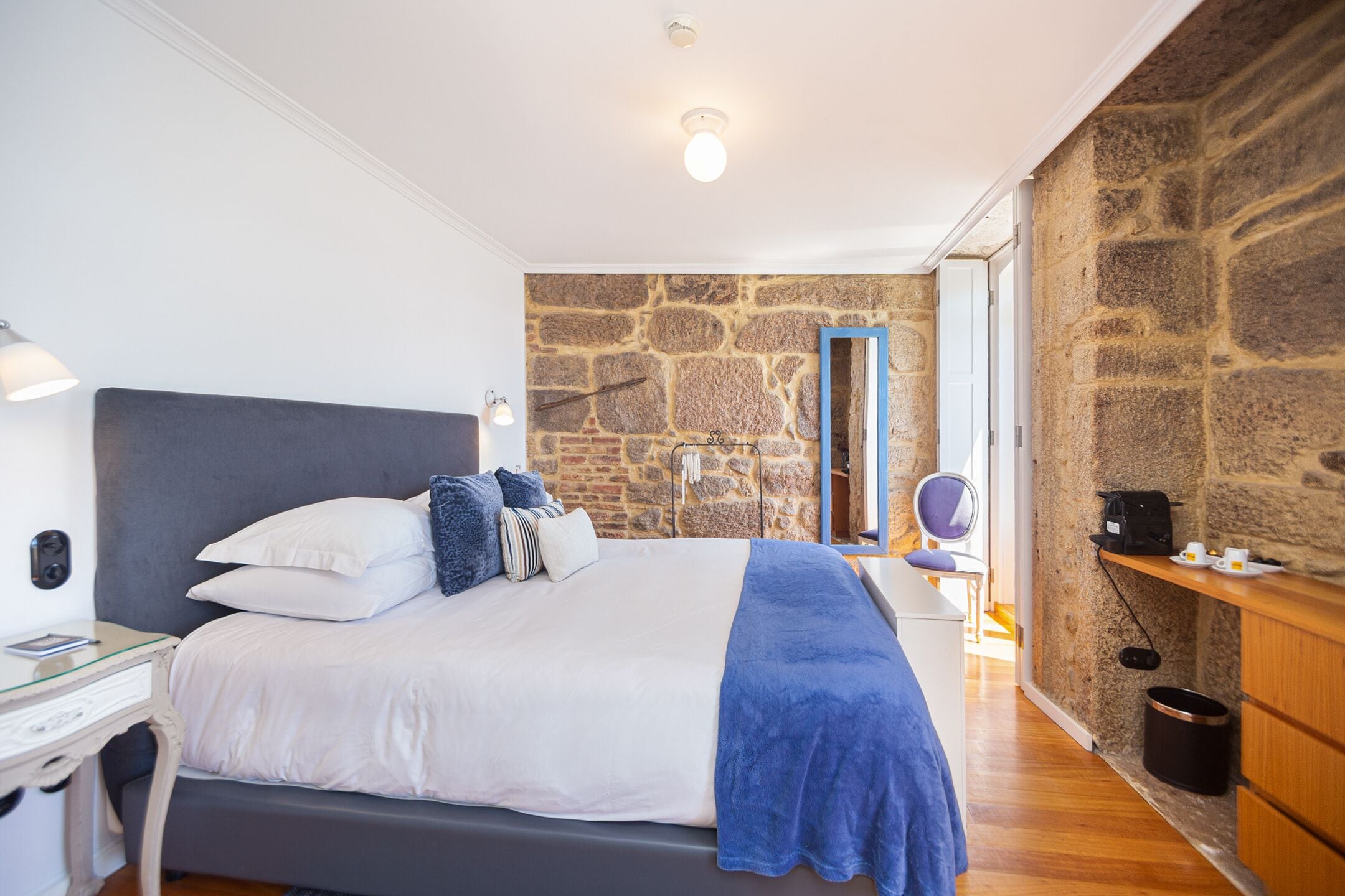 A bedroom with a large bed, a stone wall, a wooden dresser, and a desk with a lamp.