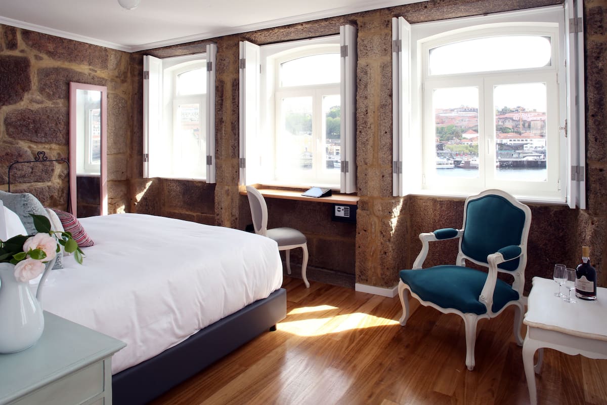 A bedroom with a large bed, a desk, a chair, and a view of a cityscape through the windows.