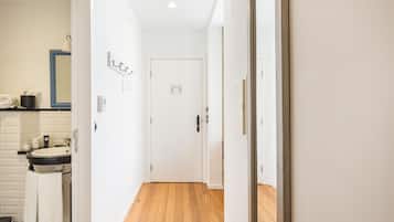 A hallway with a wooden floor leading to a white door. A bathroom with a sink and mirror is visible on the left.