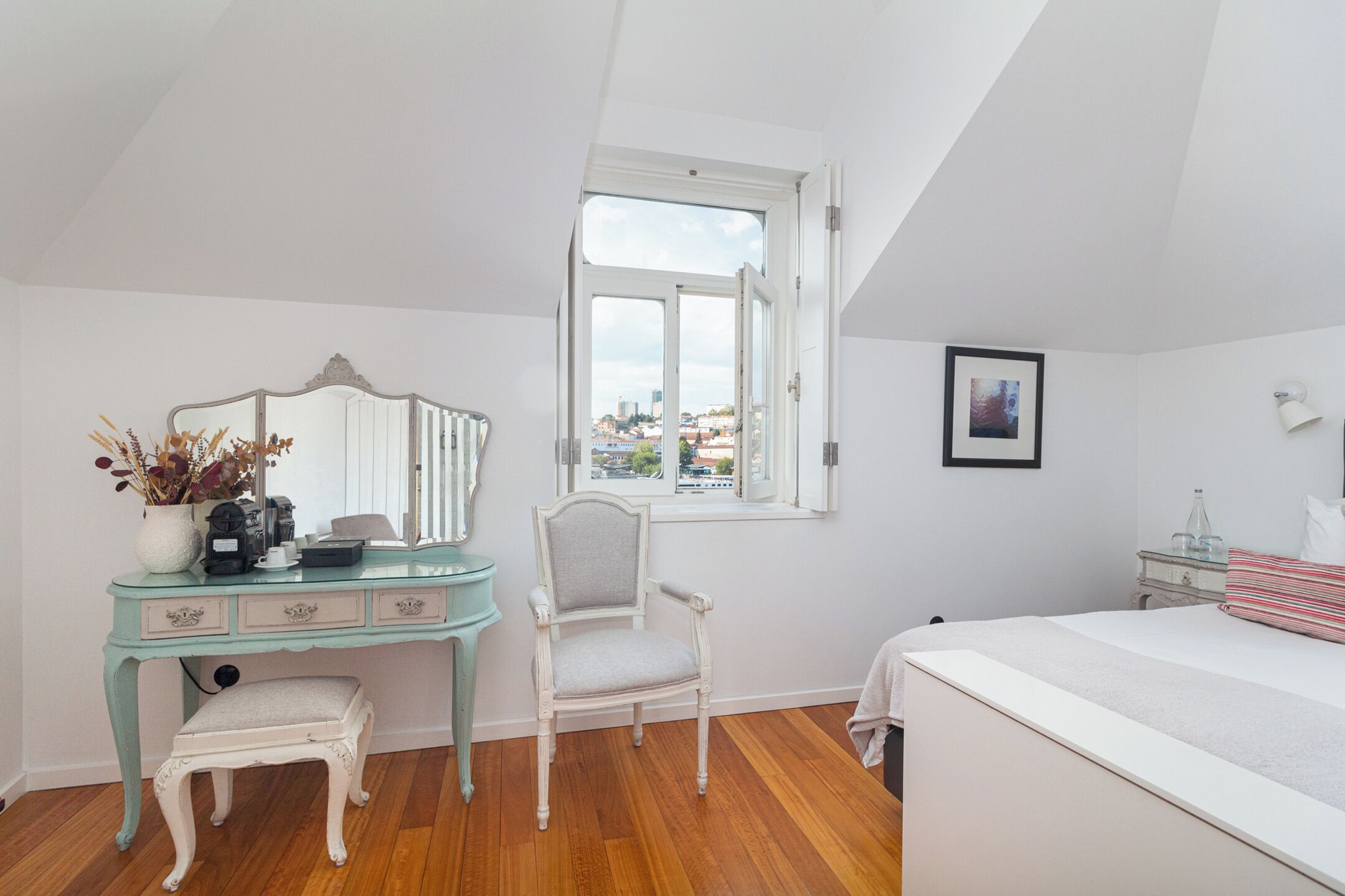 A bedroom with a bed, a vanity with a mirror, a chair, a window with a view, and a bedside table with a lamp.