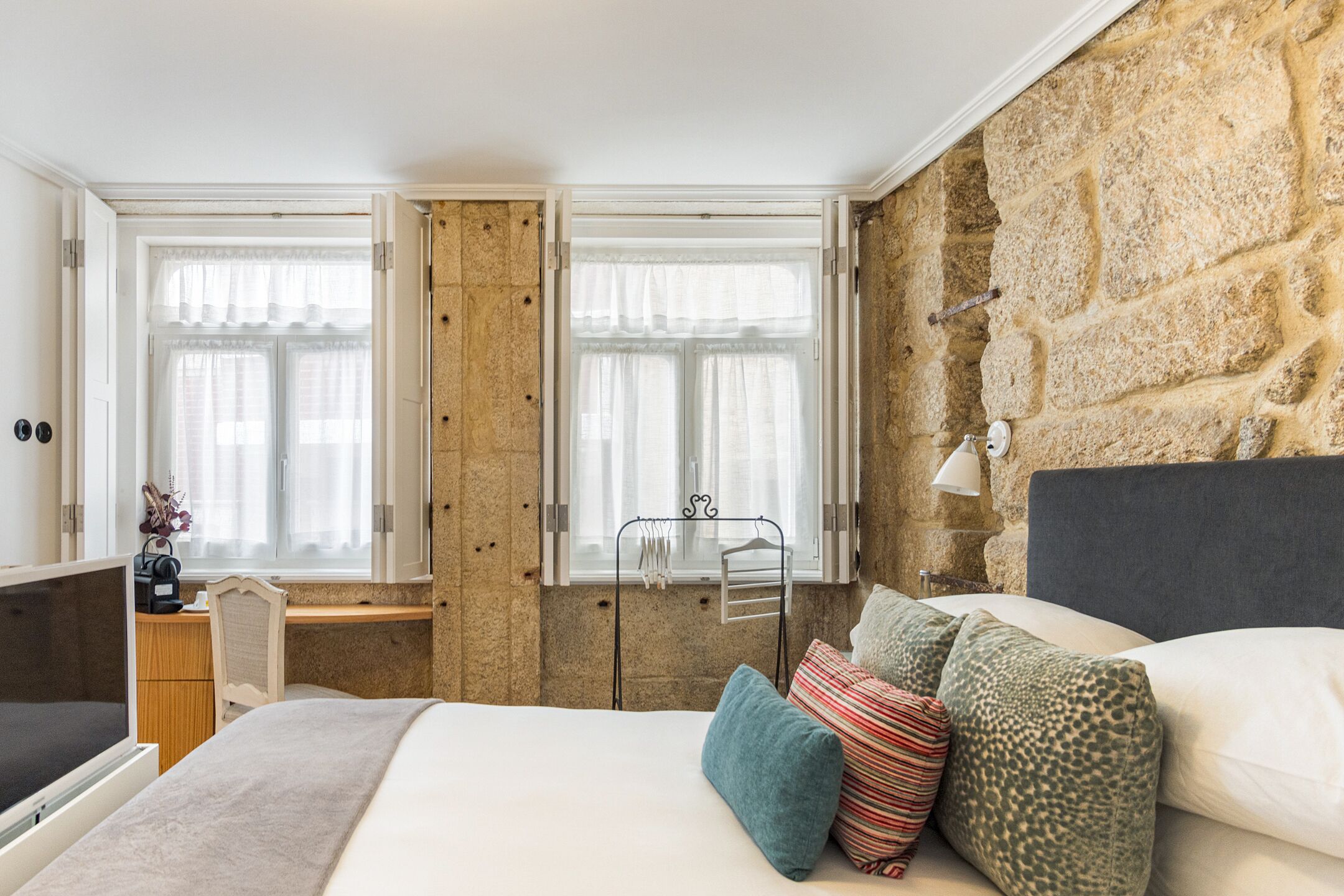 Un dormitorio con pared de piedra, cama con almohadas, escritorio con silla y ventana con cortinas.