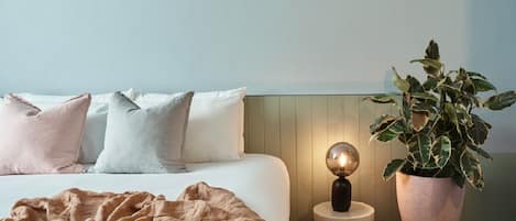 Un dormitorio con una cama, almohadas, una manta, una mesita de noche con una lámpara y una planta en maceta.