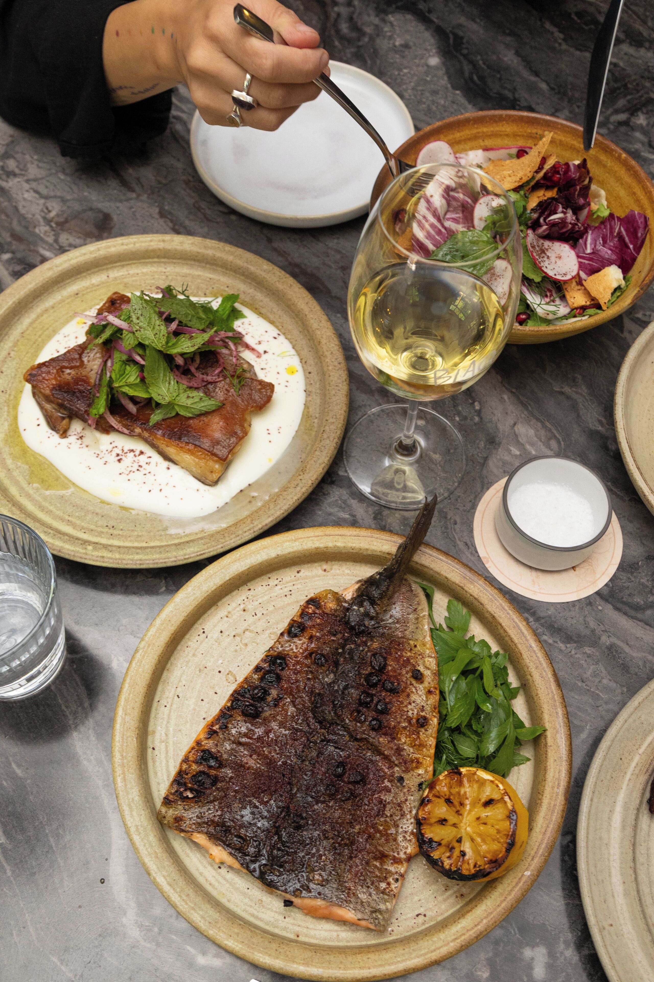 A dining table with plates of food, a glass of wine, and a person using a fork.