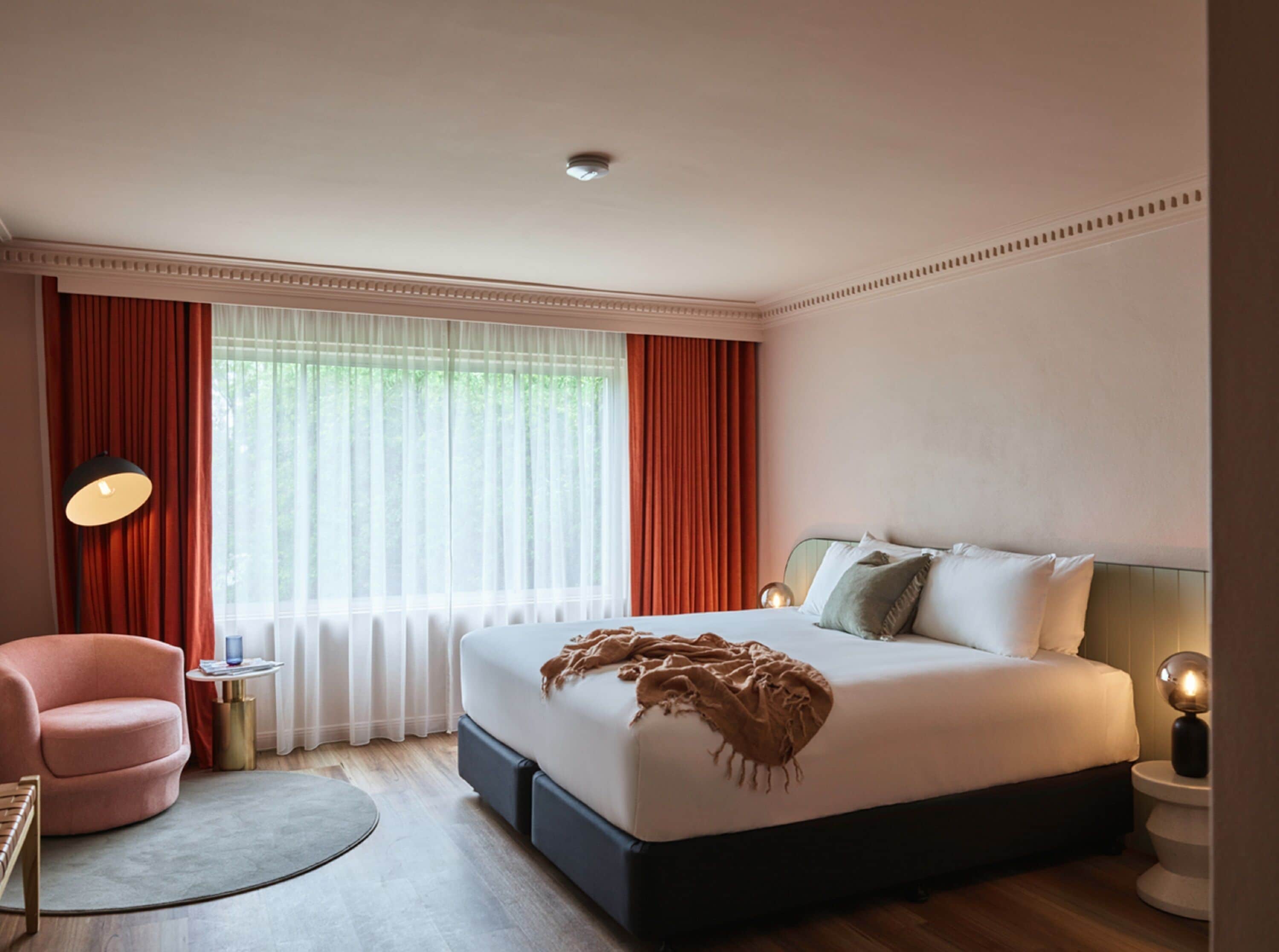 Una habitación de hotel moderna con una cama grande, una mesita redonda, una silla y un ventanal con cortinas rojas.