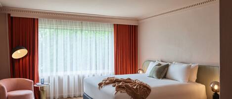 Una habitación de hotel moderna con una cama grande, una mesita redonda, una silla y un ventanal con cortinas rojas.