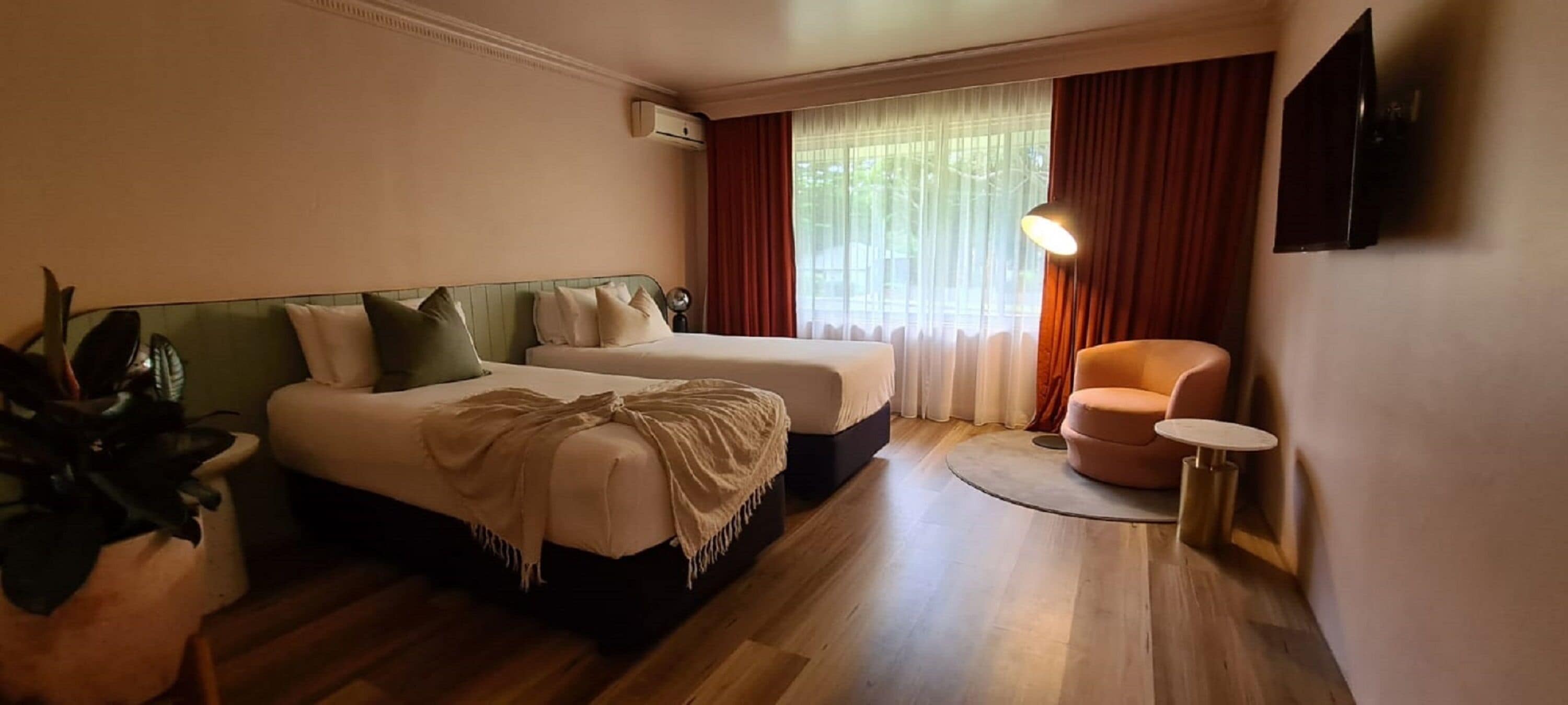 A hotel room with two beds, a small round table, a chair, a TV, and a window with curtains.