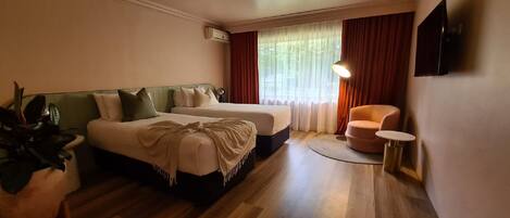 A hotel room with two beds, a small round table, a chair, a TV, and a window with curtains.