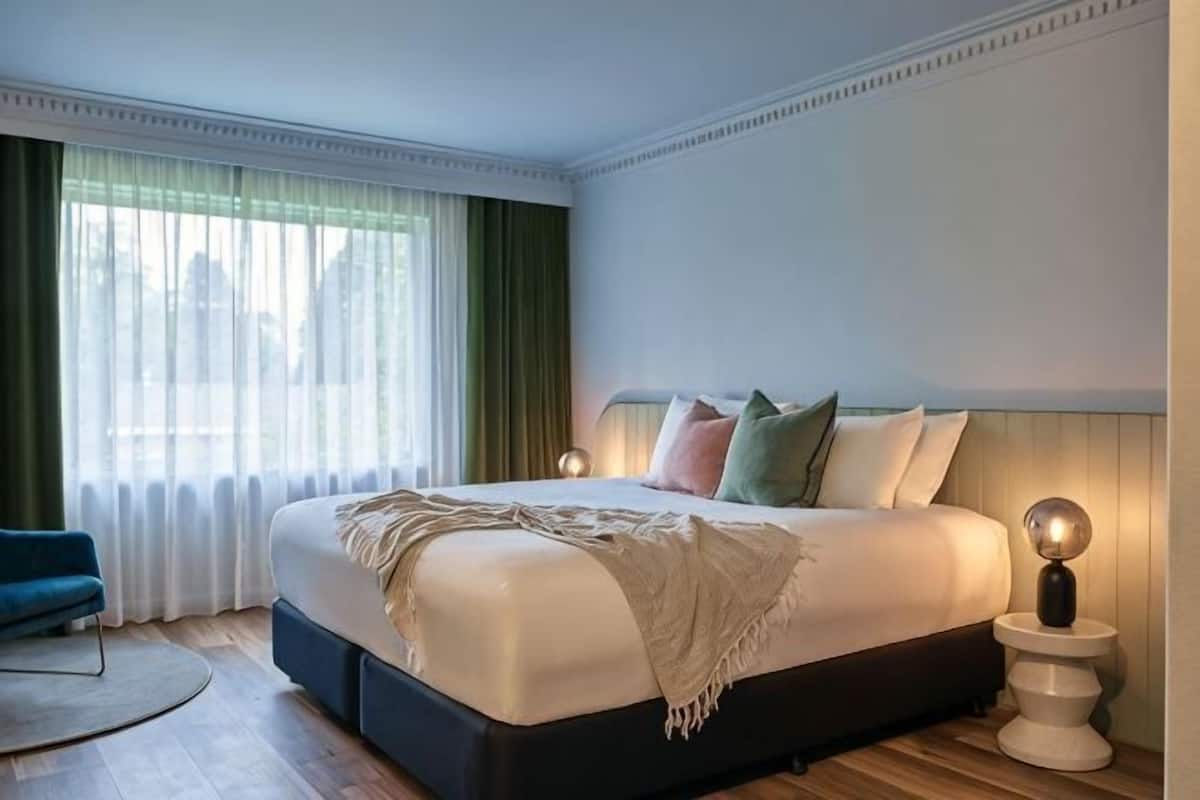 Een groot bed met hoofdeinde, een nachtkastje met lamp, een stoel en een raam met gordijnen.