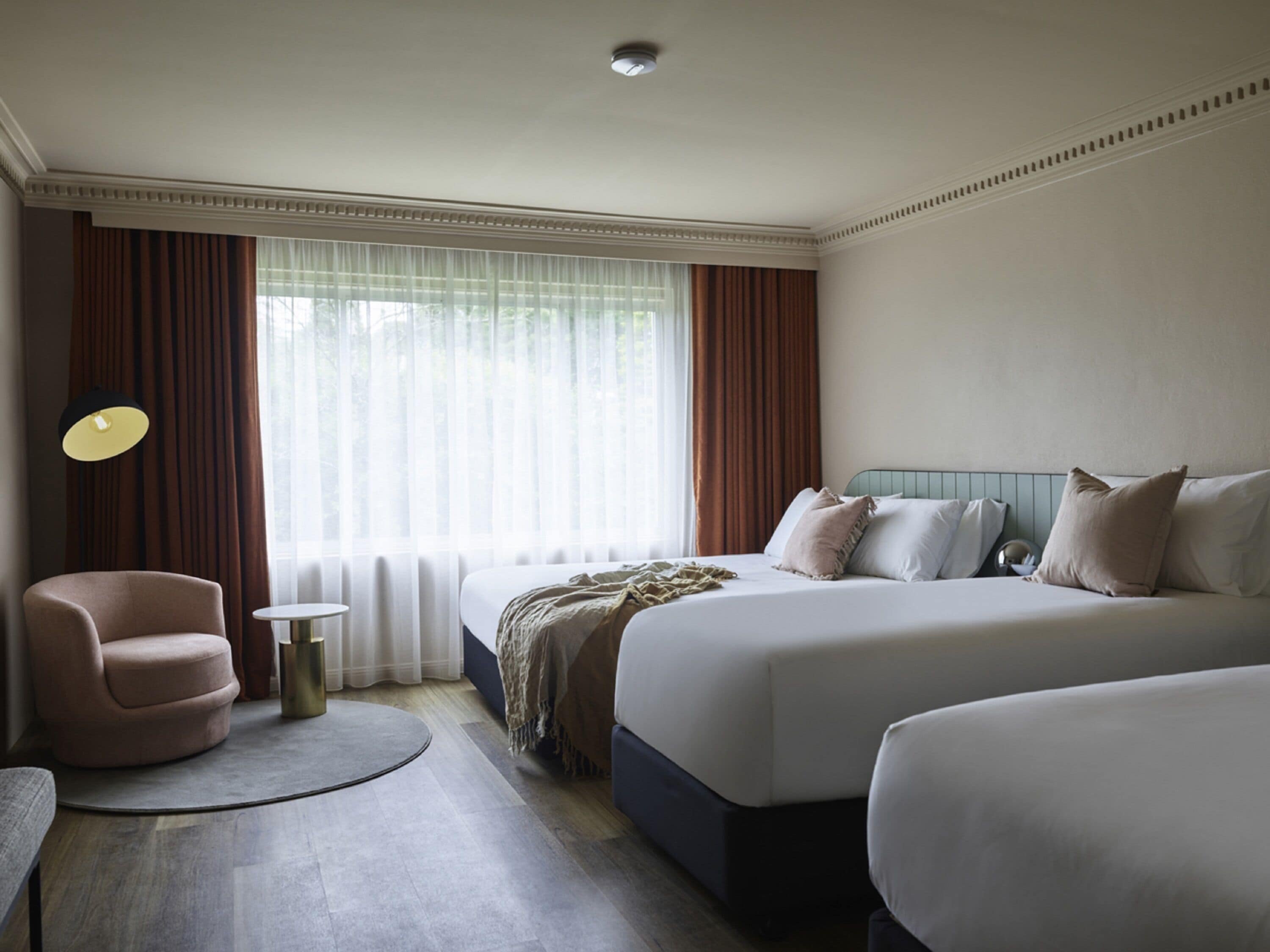 A hotel room with two beds, a chair, a round table, and a lamp.