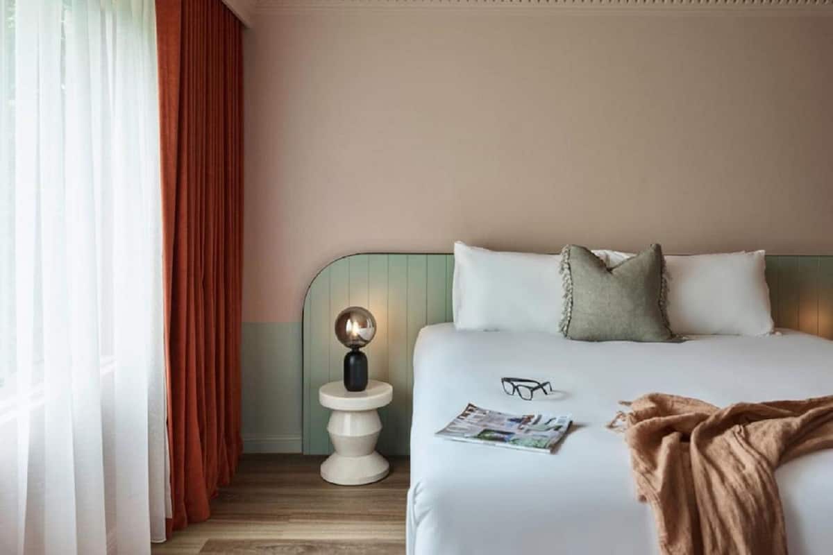Hotelkamer met een bed, nachtkastje met lamp, raam met gordijnen, en een tijdschrift op het bed.