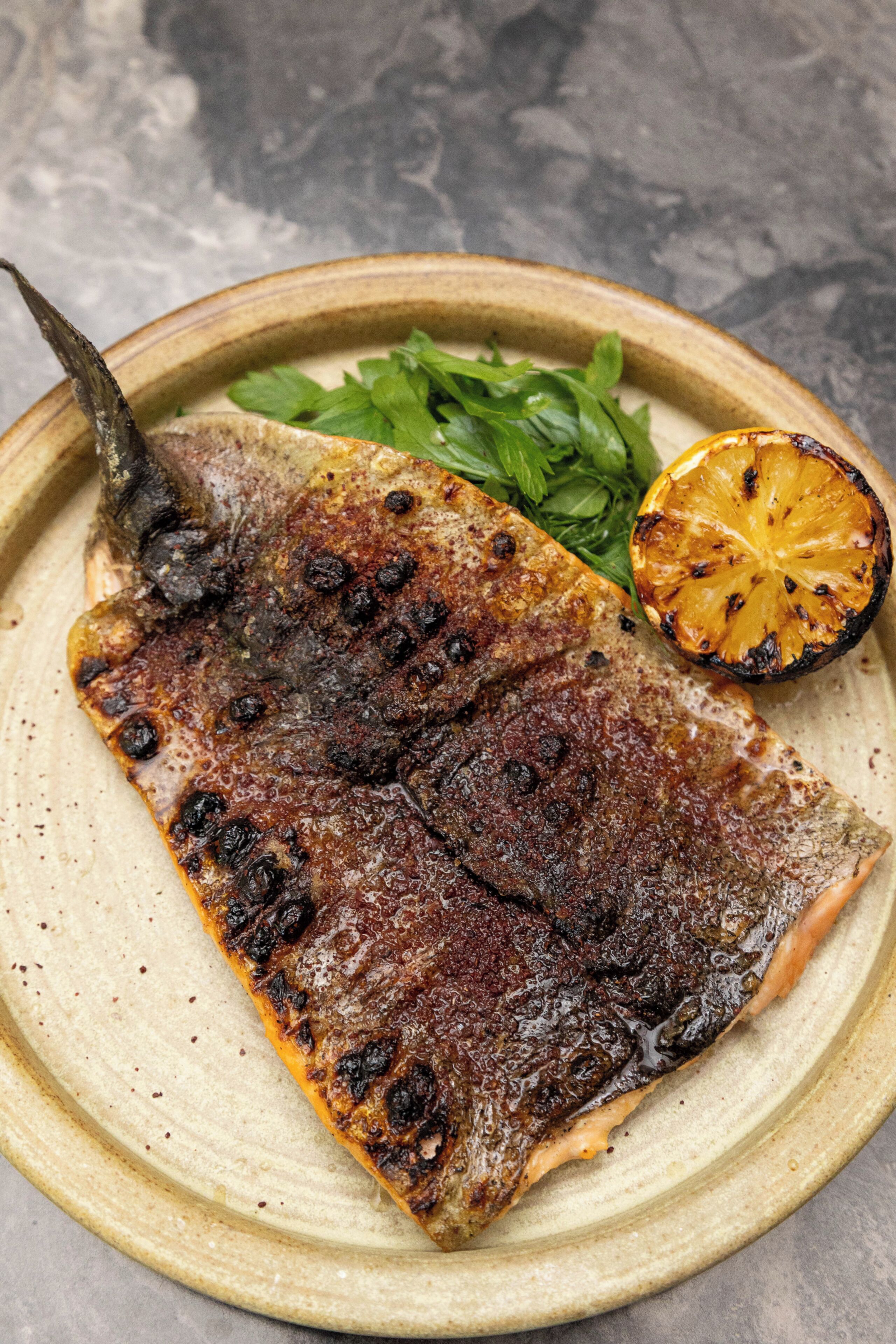 A grilled fish with a charred skin, accompanied by a slice of grilled fruit and some greens on a ceramic plate.
