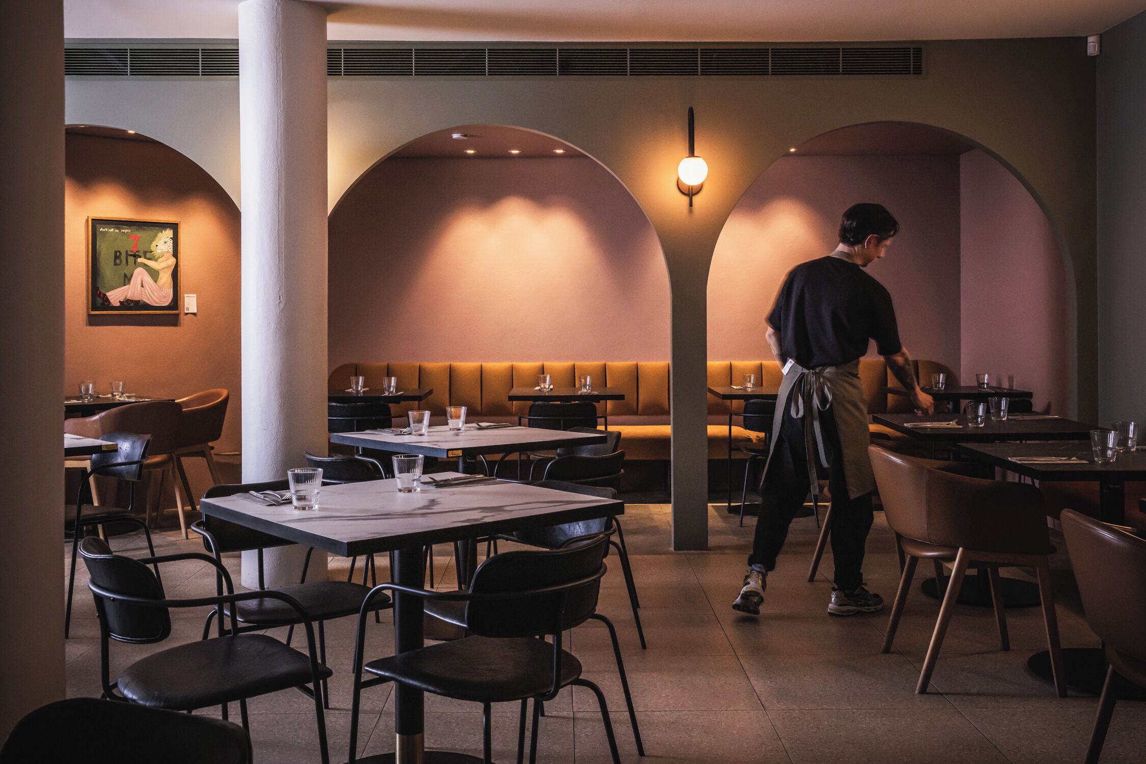 Een restaurant met tafels, stoelen, een ober en een muur met een ingelijst kunstwerk.