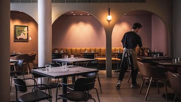 Een restaurant met tafels, stoelen, een ober en een muur met een ingelijst kunstwerk.
