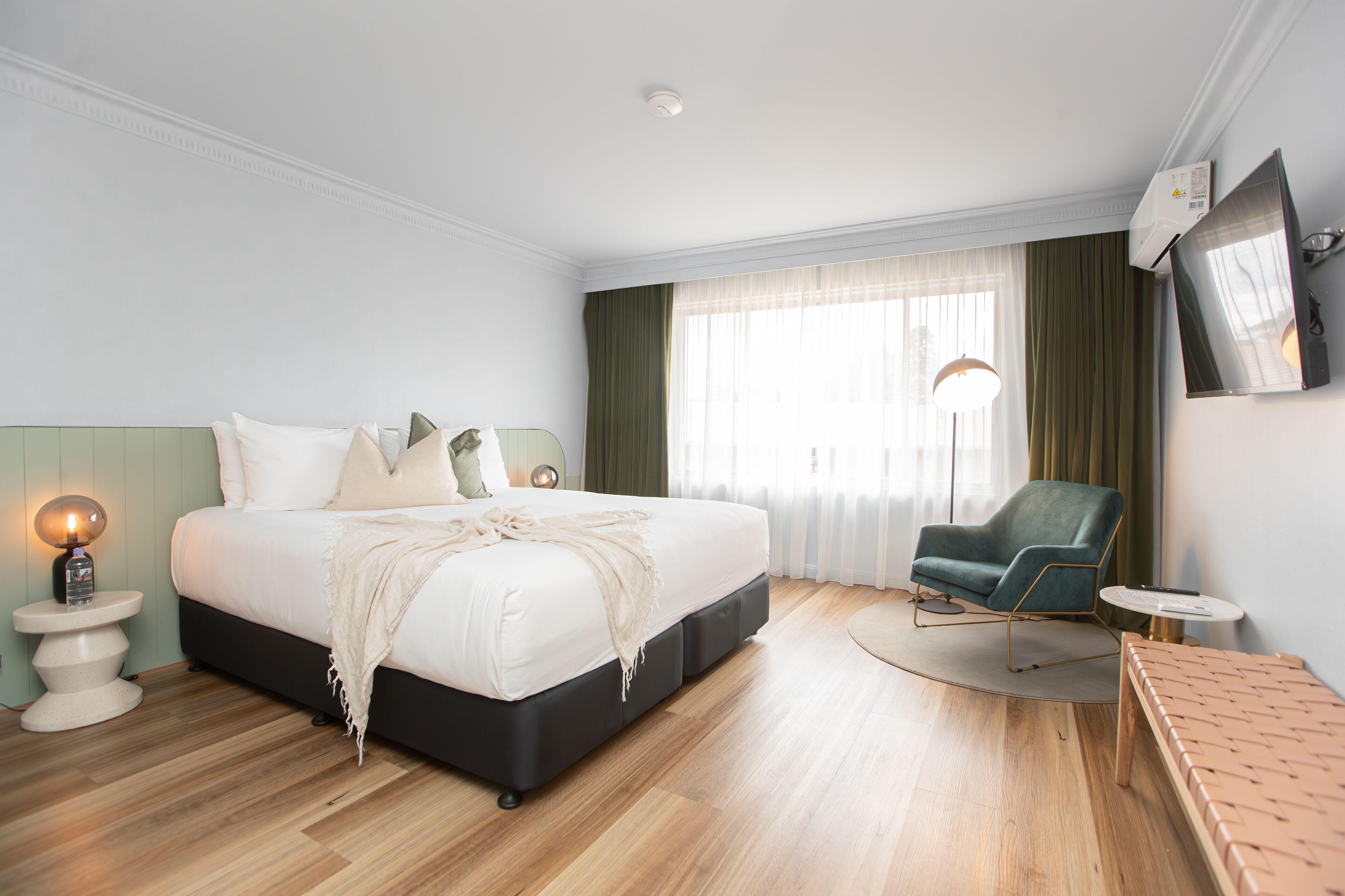 A modern hotel room with a large bed, a green armchair, a small round table, and a TV mounted on the wall.