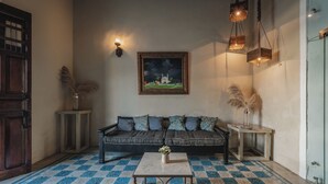 Une pièce avec un sol à carreaux, un canapé agrémenté de coussins à motifs, une petite table sur laquelle est posé un vase, un tableau accroché au mur et des lampes suspendues.