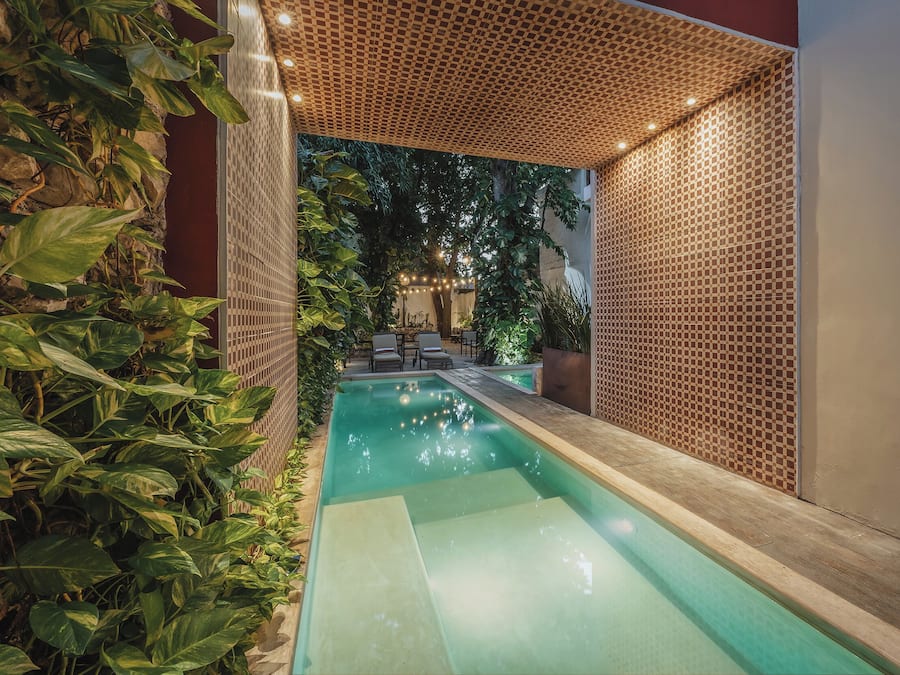 Una piscina con cubierta de madera, rodeada de abundante vegetación y un muro decorado.