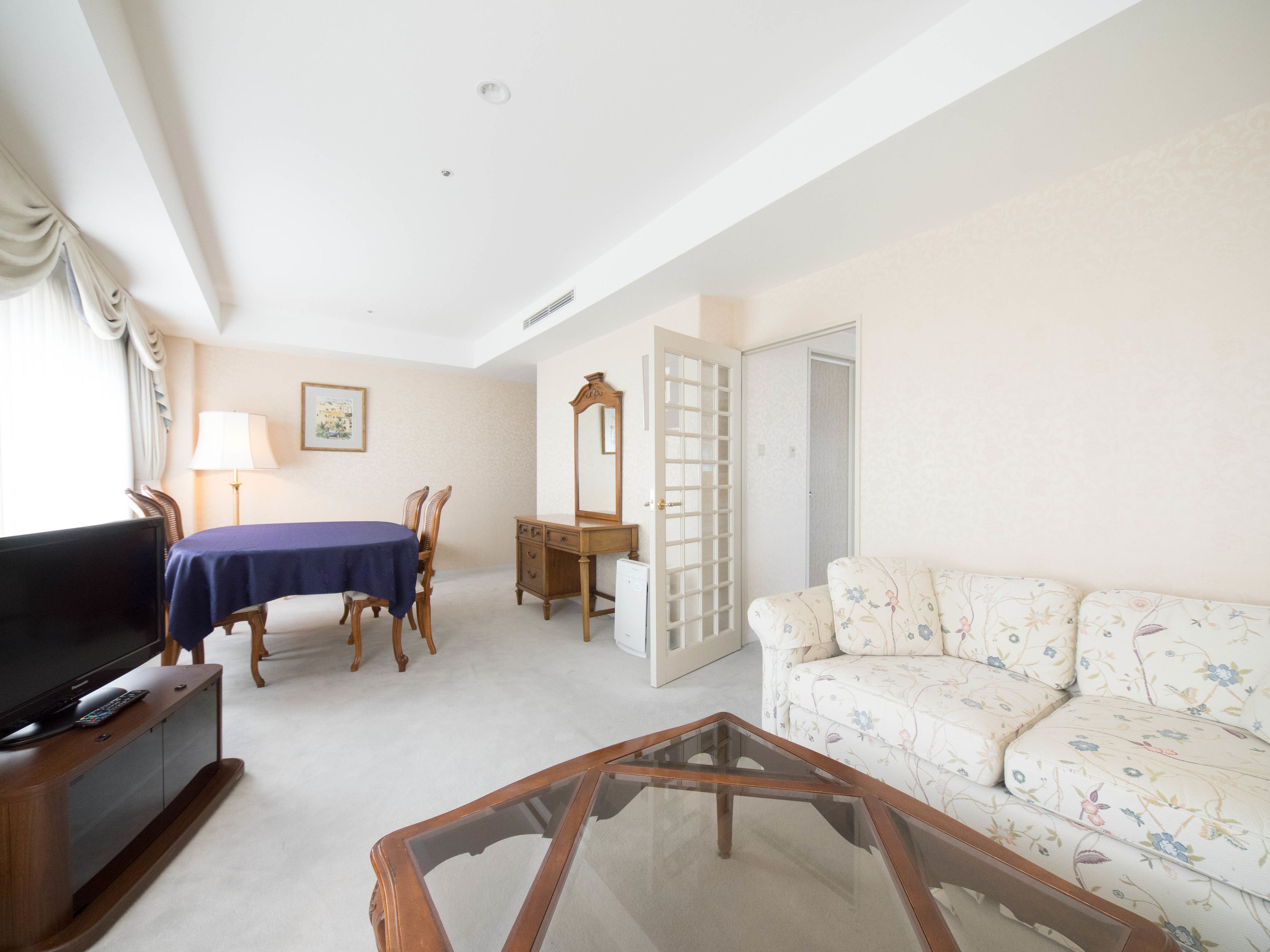 Un salon comprenant un téléviseur, une table basse en verre, un canapé à motifs floraux, une table à manger avec des chaises, une commode en bois et une porte donnant sur une autre pièce.