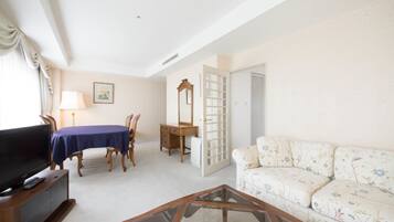 Un salon comprenant un téléviseur, une table basse en verre, un canapé à motifs floraux, une table à manger avec des chaises, une commode en bois et une porte donnant sur une autre pièce.